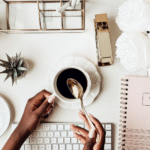 An African American young girl hand stirring coffee on a desk, with a rose gold spiral notebook, a stapler and a pen holder.
